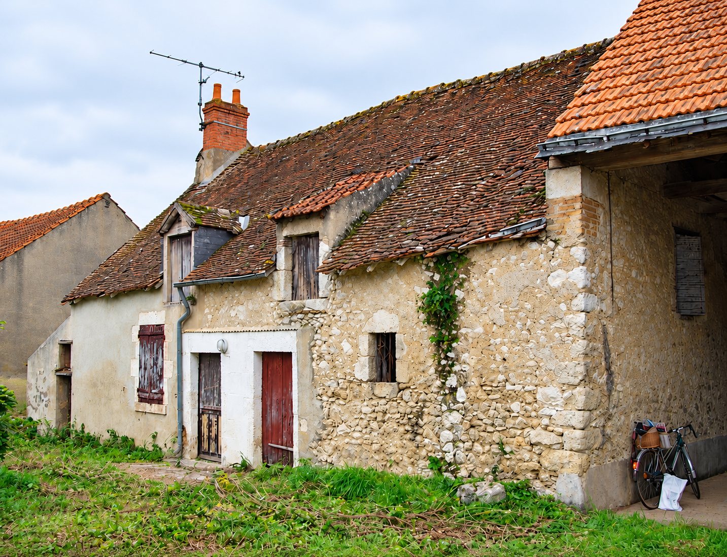 Une vieille maison en pierre au toit de tuiles brunes, avec des volets usés, une porte rouge et un vélo garé sous l'auvent d'une grange.