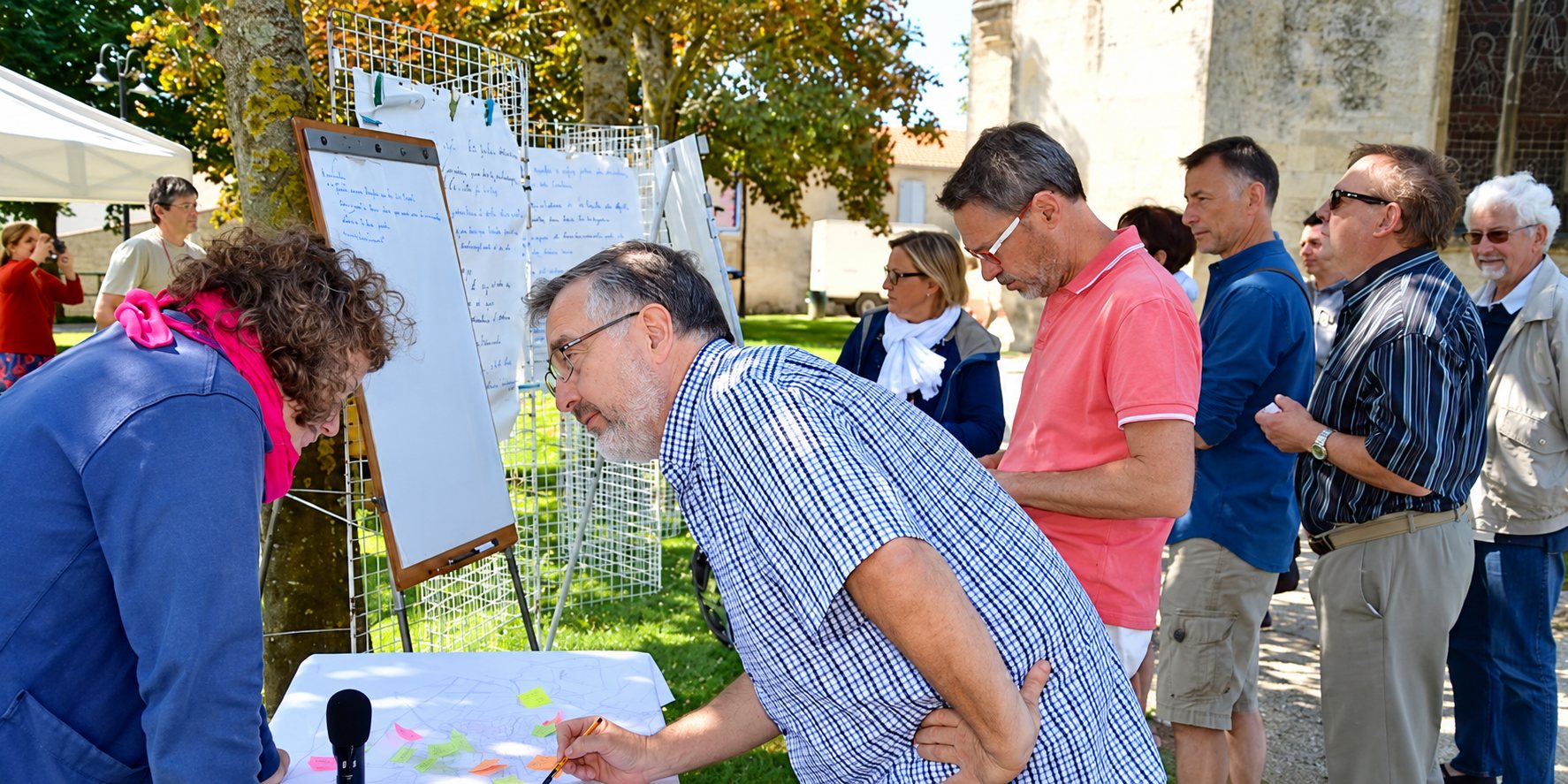 Des hommes et des femmes se rassemblent en extérieur autour d'une table et de panneaux d'affichage lors d'un atelier participatif, près d'une église.