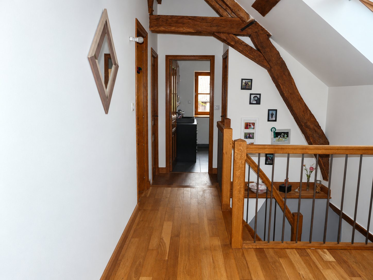 Un couloir sous les combles avec parquet en chêne, poutre en bois apparente, garde-corps à barreaux métalliques et porte ouverte sur une salle de bain.