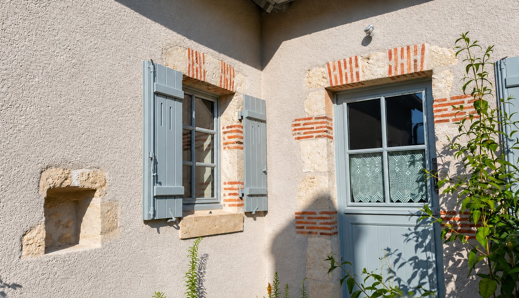 Façade crépie d'une maison ancienne avec fenêtre à volets gris-bleu ouverts, porte vitrée assortie et encadrements en pierre et briques.