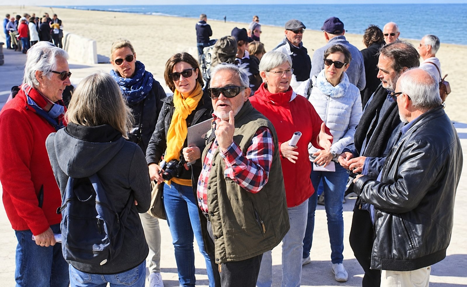Un groupe d'adultes en vêtements chauds est réuni en plein air sur une promenade au bord de la mer et écoute une personne qui parle en levant le doigt.
