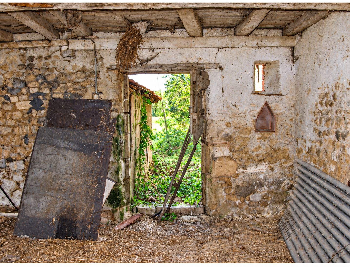Intérieur d'une vieille grange en pierres avec poutres apparentes, paille au sol et une porte ouverte sur la végétation.