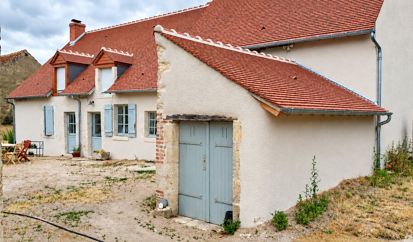Maison de campagne rénovée à la façade enduite claire, au toit de tuiles rouges et aux volets gris-bleu, avec une petite annexe à porte en bois.