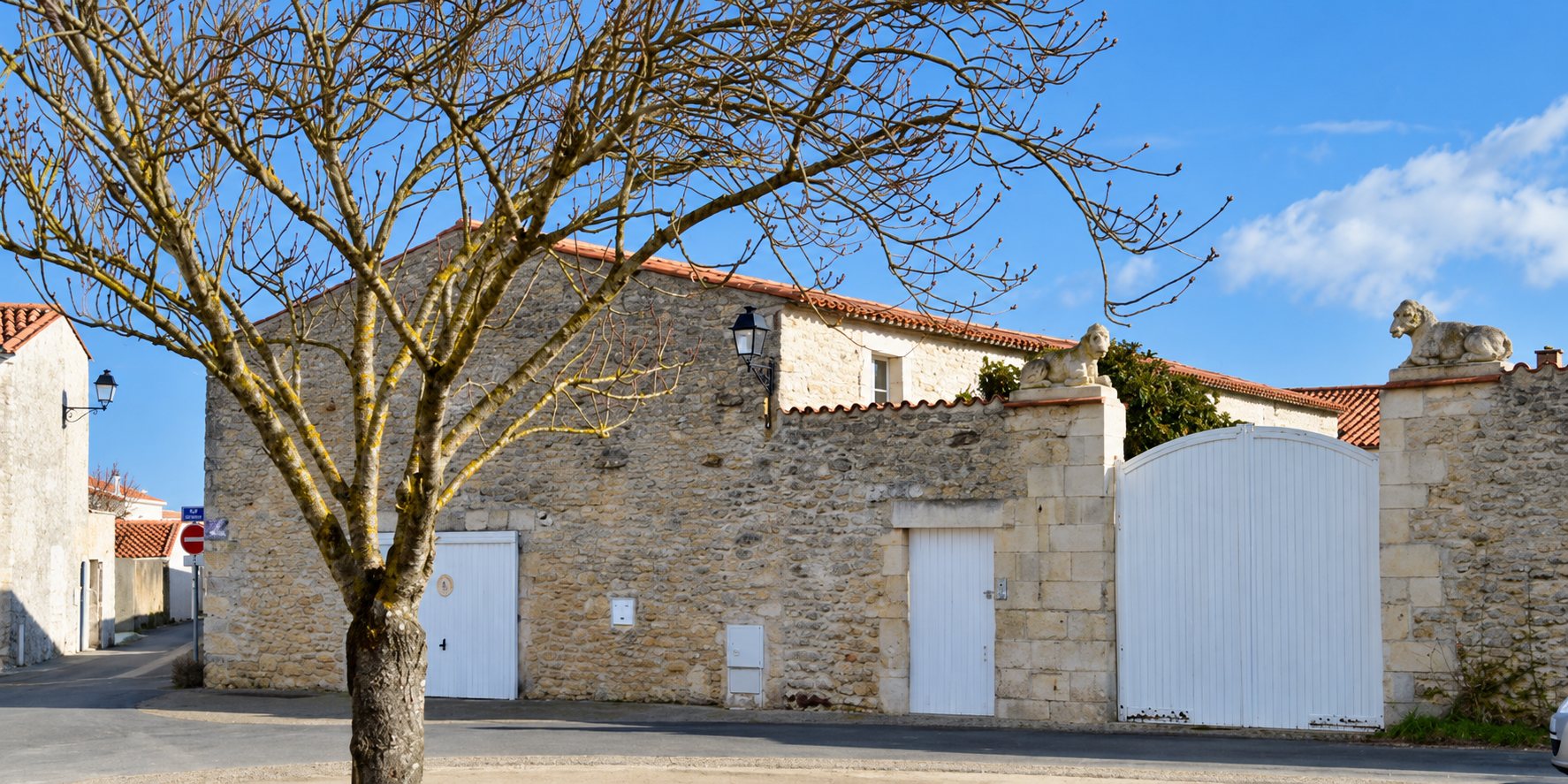 Une maison en pierre au toit de tuiles rouges, dotée d'un grand portail blanc et surmontée de statues de lions, derrière un arbre sans feuilles.