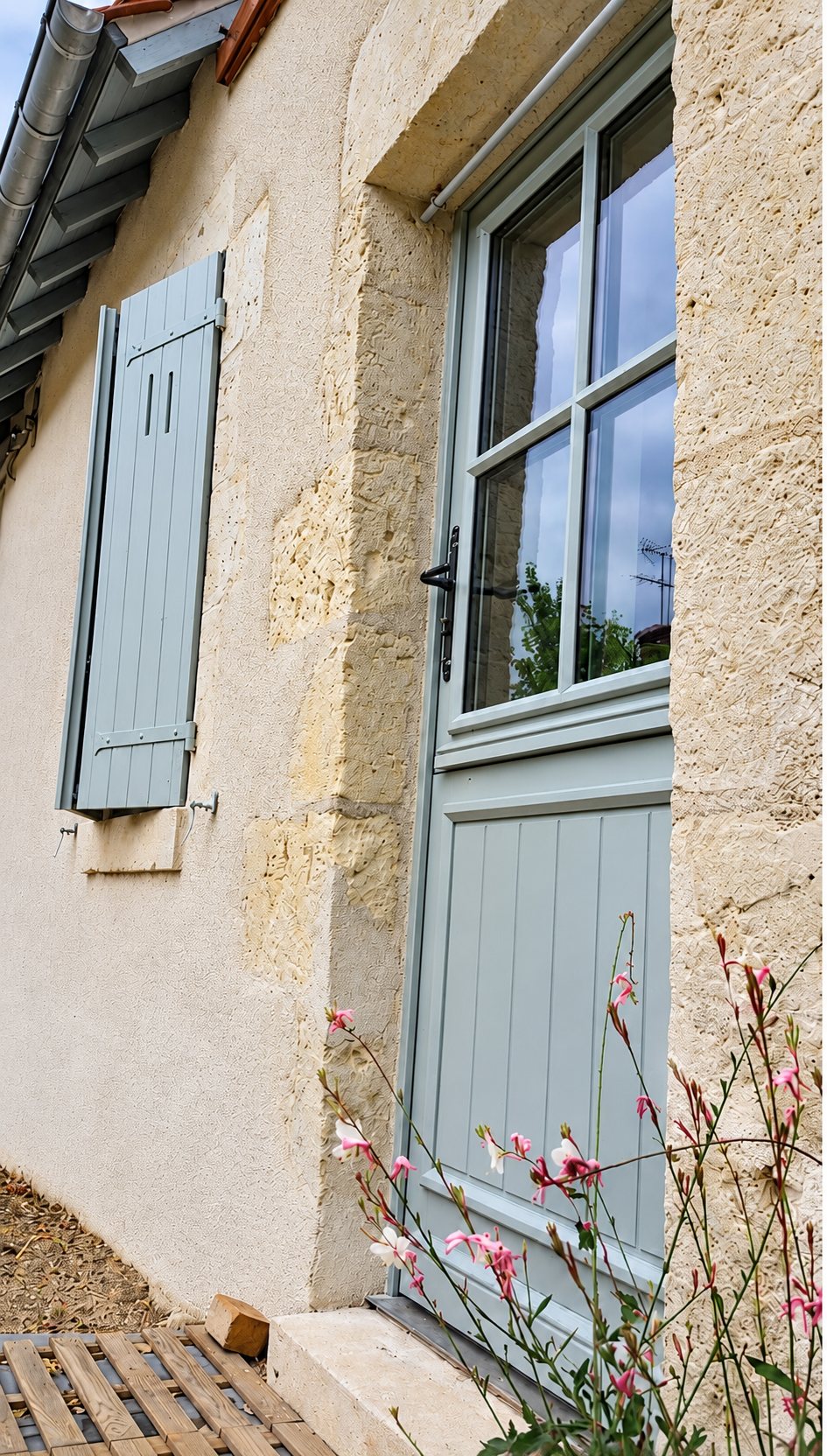 Façade en pierre claire d'une maison avec une porte d'entrée vitrée et un volet en bois peints en gris-vert, des fleurs roses au premier plan.