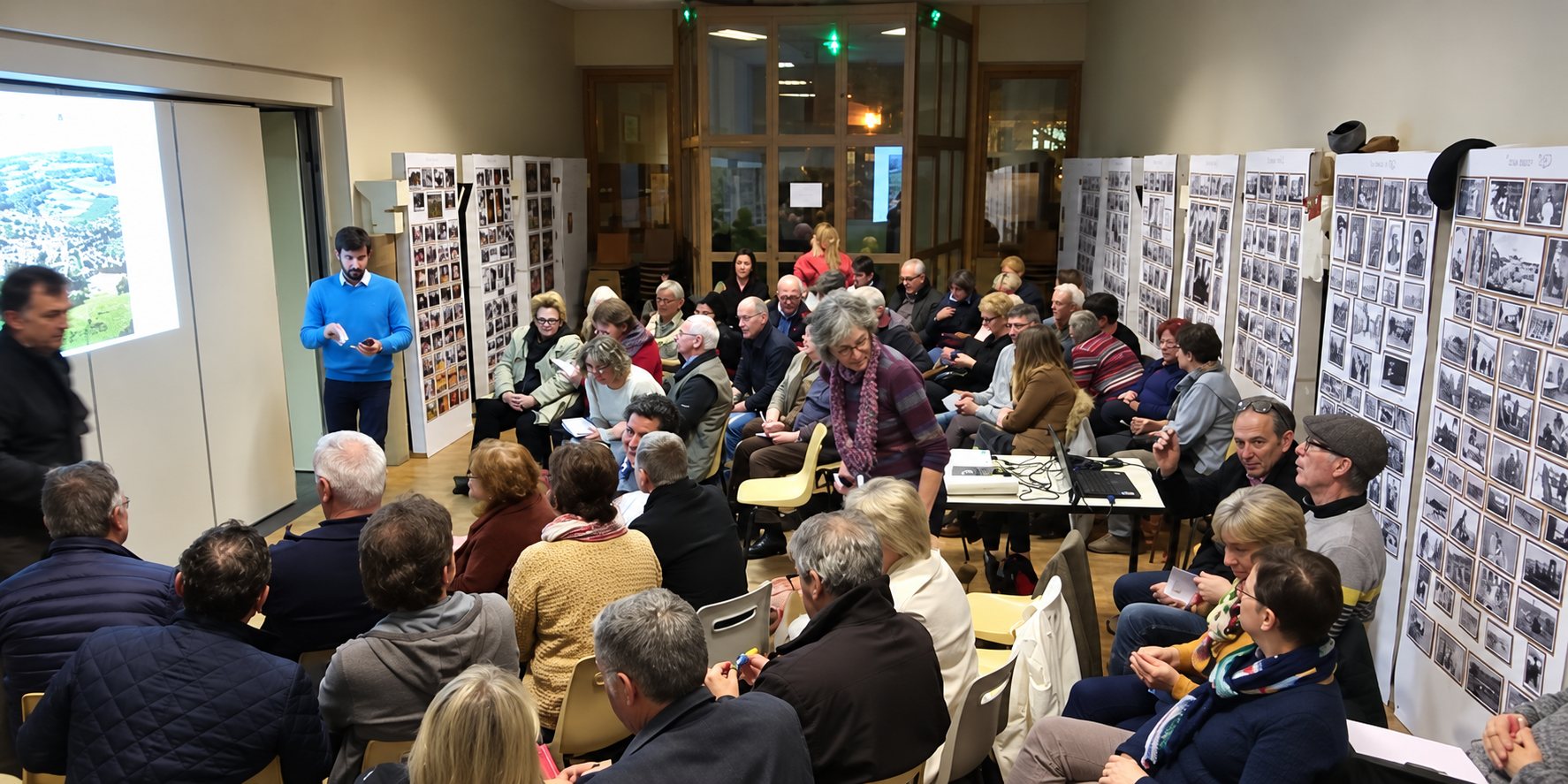 Une foule de personnes assises dans une salle assiste à une présentation projetée à l'écran, entourée de panneaux couverts de photographies anciennes en noir et blanc.