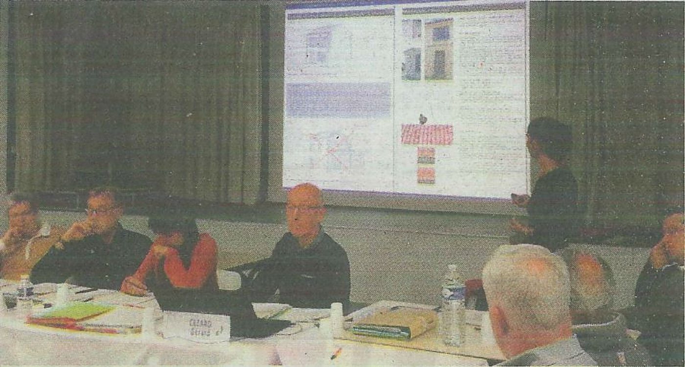 Des personnes assises autour d'une table lors d'une réunion, tandis qu'une personne debout présente un document projeté sur un grand écran.