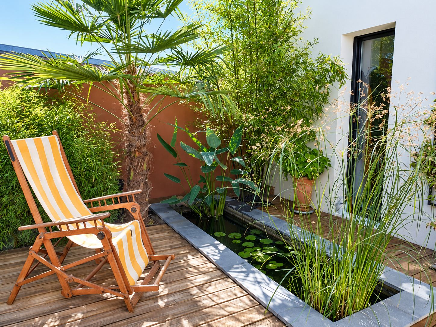 Une terrasse en bois ensoleillée aménagée avec un petit bassin rectangulaire bordé de pierre, un palmier, des bambous, des graminées et une chaise longue à rayures jaunes et blanches.