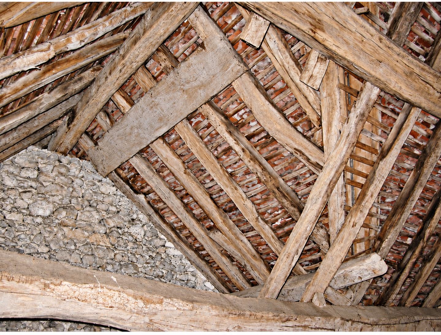 Vue de dessous d'une ancienne charpente en bois brut supportant une toiture de tuiles en terre cuite, avec un mur de pierres à gauche.